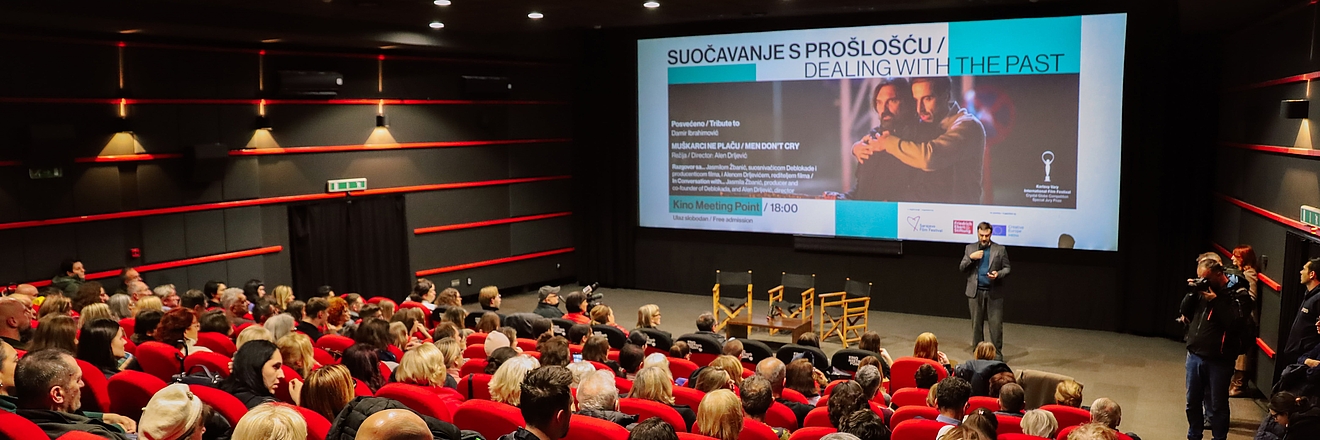 A full cinema auditorium with rows of red seats filled with people watching a speaker on stage. The speaker stands in front of a large screen displaying the title “Suočavanje s prošlošću / Dealing with the Past,” along with event details and an image from a film. Several empty director-style chairs are set up on stage, and a camera crew is positioned on the right side of the room.