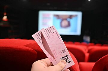 Two film tickets being held in empty cinema in front of banner announcing an upcoming film. 